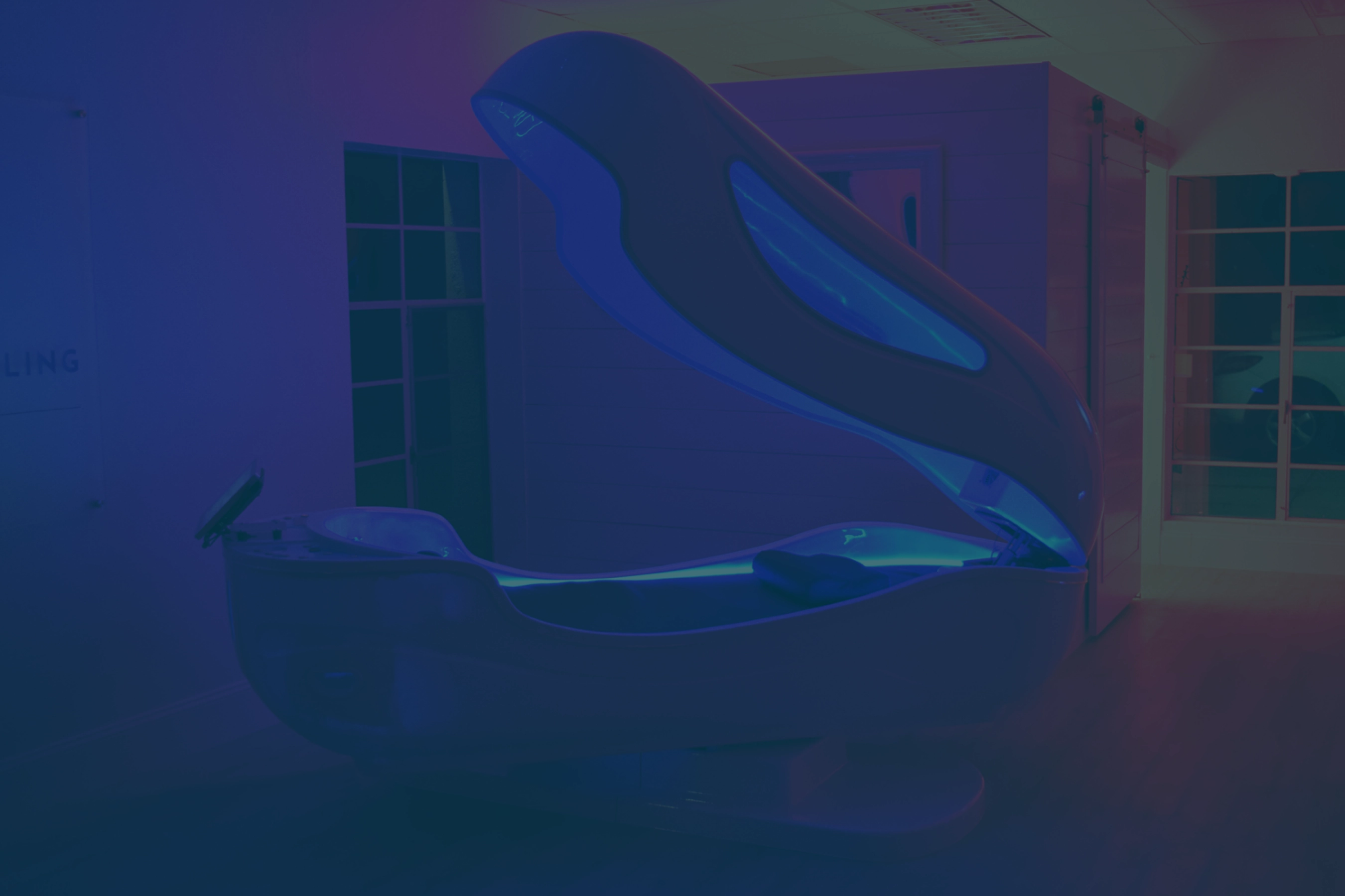 An open, modern floatation therapy tank with glowing blue interior light sits in a dim room with a window and a sliding barn door.