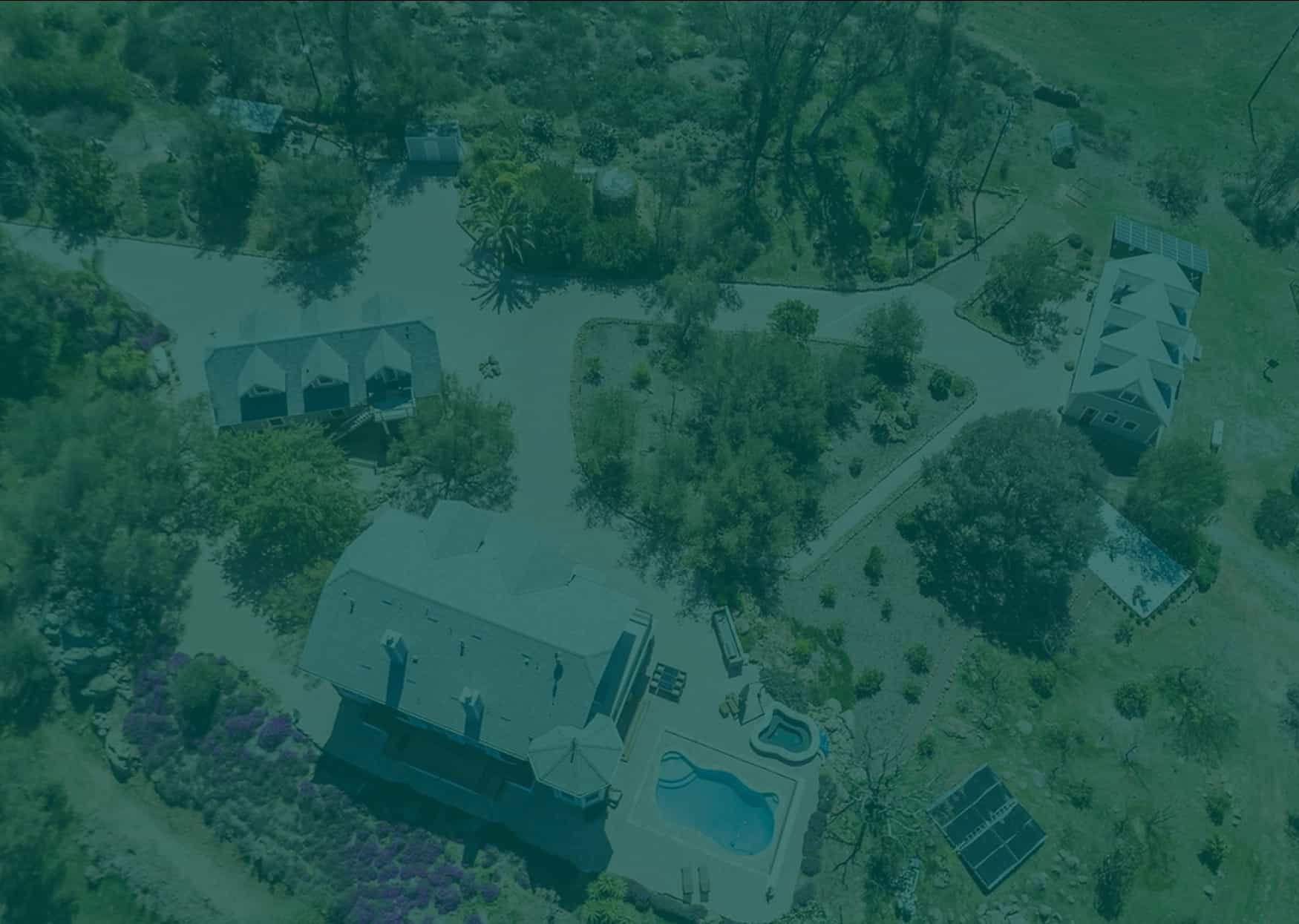 Aerial view of a large rural estate featuring multiple houses, winding driveways, a swimming pool, hot tub, and several solar panel arrays, all surrounded by green trees and fields.