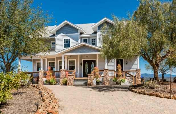 A light grey, two-story modern farmhouse-style house with a covered front porch, stone pillars, and a paved driveway. Large trees frame the house under a clear blue sky.