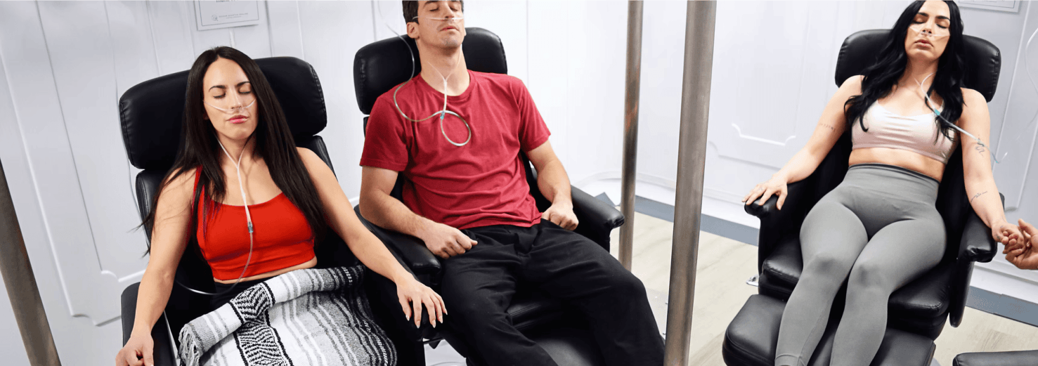 Three people, two women and one man, sit in black chairs receiving oxygen via nasal cannulae. They appear relaxed, and one woman holds hands with an unseen person.