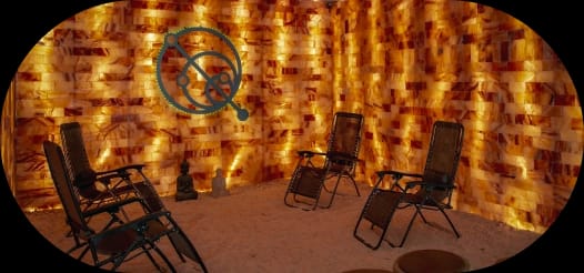 An illuminated salt room featuring walls of glowing salt bricks, a salt-covered floor, and several recliner chairs arranged for relaxation.