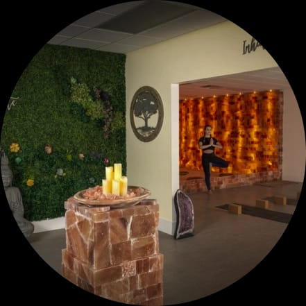 A serene wellness studio featuring a woman doing yoga in a glowing salt therapy room, a green living wall, a salt pillar with candles, and a Buddha statue.