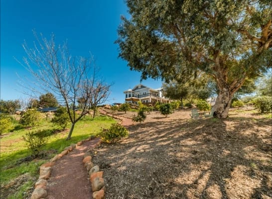A large, multi-story house on a hill, accessed by a winding path, with a spacious lawn and a large, leafy tree shading two Adirondack chairs in the foreground, under a bright blue sky.