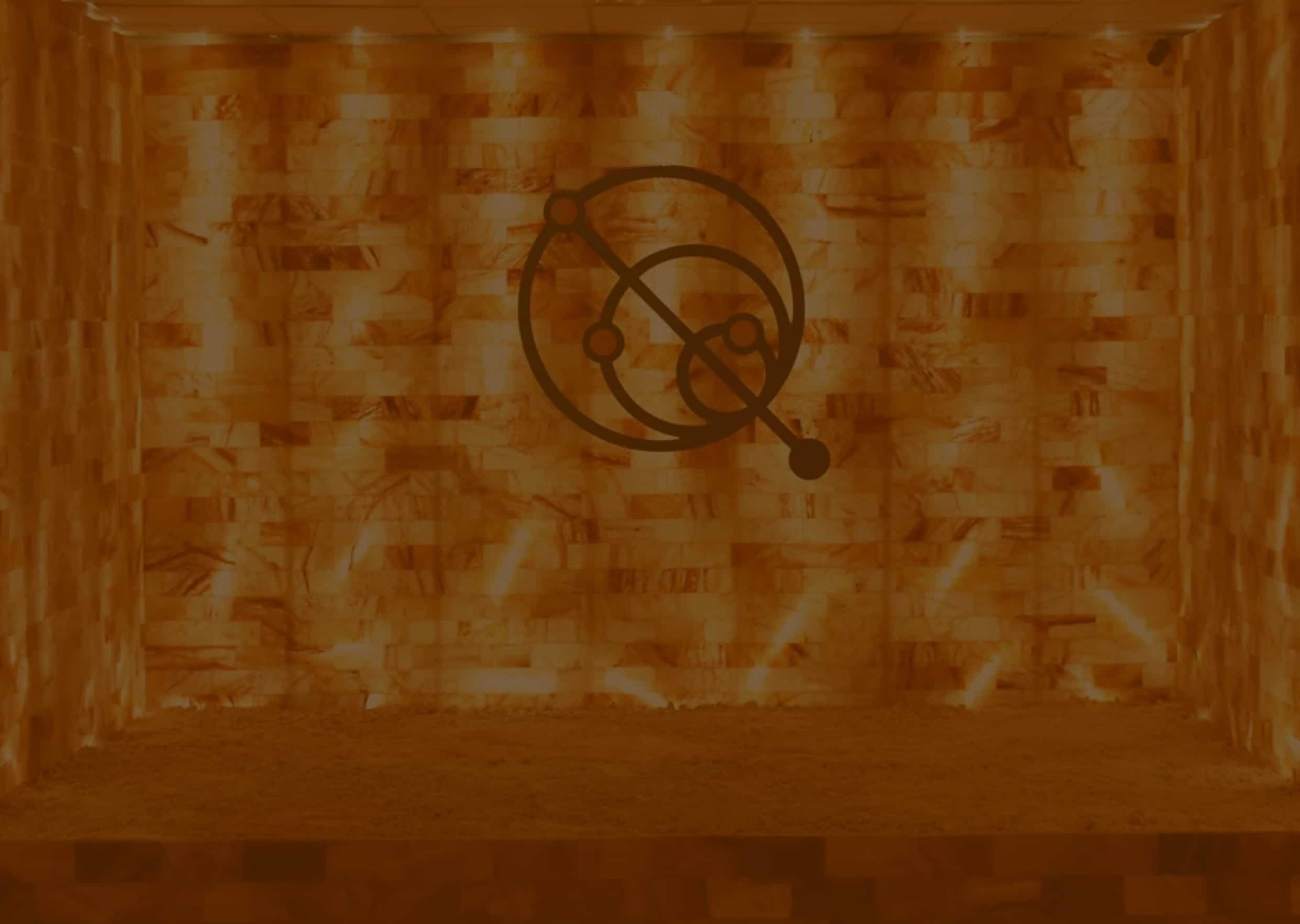 Interior of a warm orange salt room with glowing brick walls, a sandy floor, and a dark abstract circular logo in the center.