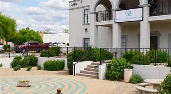 Exterior of the Quasar Quantum Healing clinic building, featuring stairs to the entrance and a gravel courtyard with a blue labyrinth pattern.
