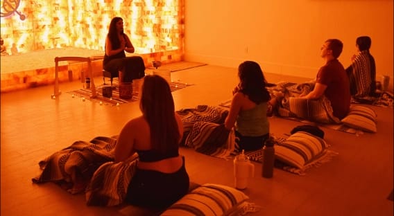 A group meditates on mats and cushions in a warm, dimly lit room with an illuminated salt crystal wall behind the seated instructor.