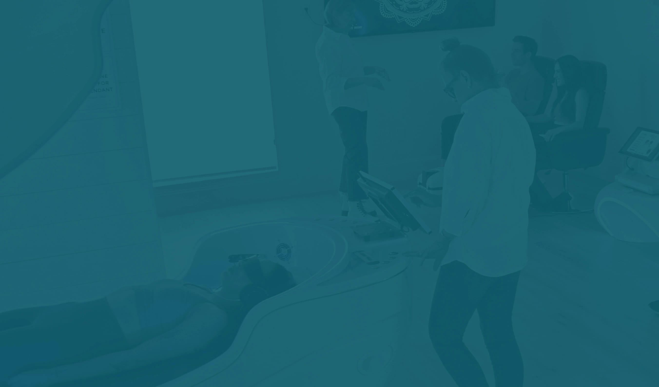 A person lies in a high-tech wellness pod while a technician operates a control panel, with two people seated in the background of the modern facility.