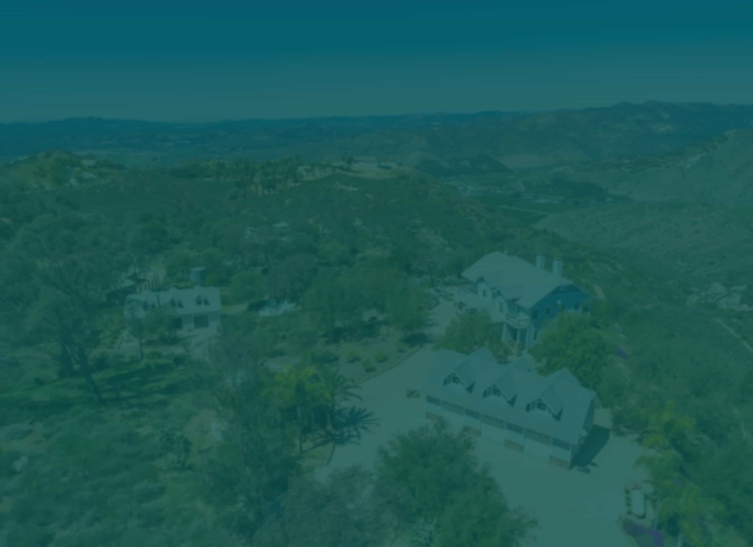Aerial shot of a large, beautiful property with multiple buildings, lush trees, and rolling green hills under a clear blue sky.